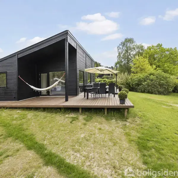 Sort, moderne hus med en hængekøje på terrassen; derudover står et havemøbelsæt med parasol. Omgivet af en grøn have med buske og træer under en blå himmel.