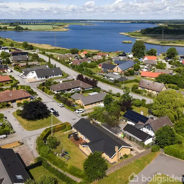 Boligområde med huse omgivet af grønne haver, set fra oven. I baggrunden ses en blå sø og åbne marker under en delvis skyet himmel.
