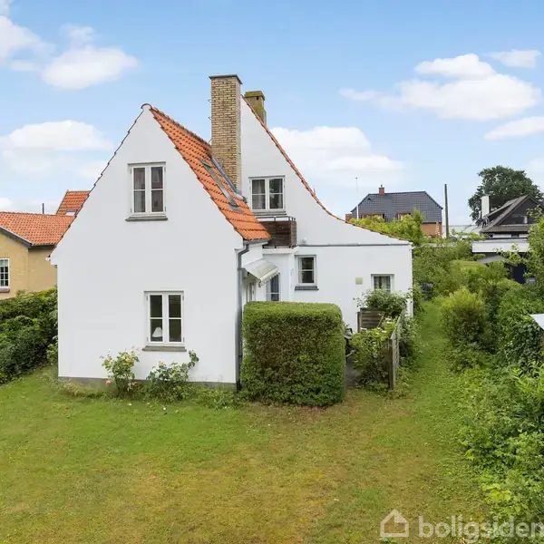 Et hvidt hus med rødt tegltag står i en velplejet have omgivet af hække. Det ligger i et beboelsesområde med flere huse og grønne områder.