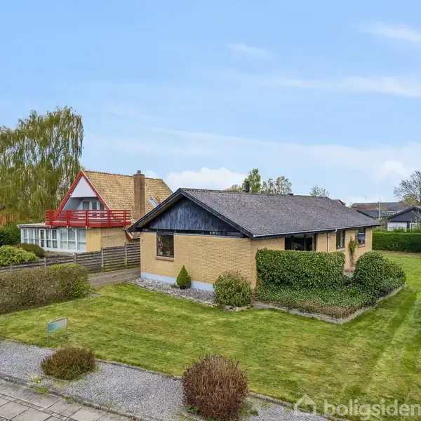 Et enfamiliehus med fladt tag står på en grøn græsplæne omgivet af hække. Baggrunden har flere træer og et nabohus med skråtag. Himlen er klar og blå.