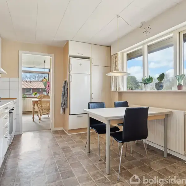 Et lyst køkken med hvidt interiør, hvor et bord med fire stole står ved vinduet. Køleskabet er indbygget i skabsvæggen, og en dør fører til en spisestue.