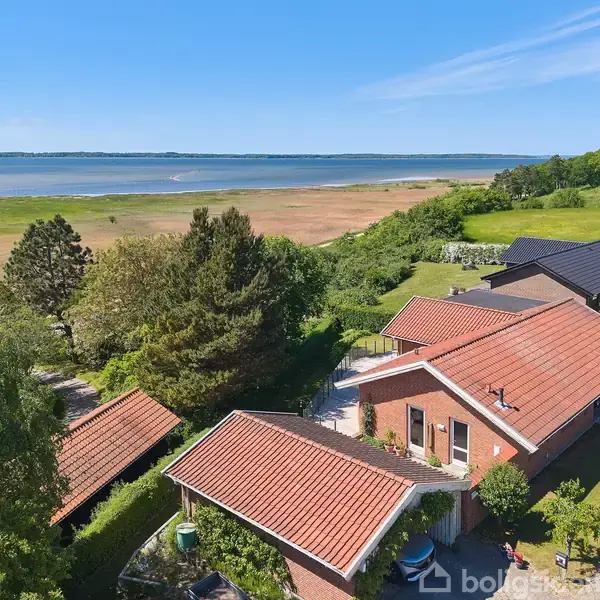 Rødt murstenshus med saddeltag ses fra oven, placeret i et grønt, kystnært område. Flere træer omgiver huset, der ligger tæt på en strandeng og farvande.