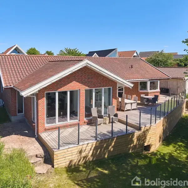 Rødt murstenshus med højt tag står stille. Stor terrasse med glasrækværk omgiver det, møbleret med bord og stole. Grønt græs og buske i villakvarter.