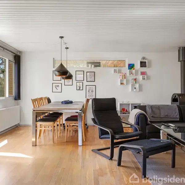 Stue med spisebord og stole til venstre, en sofa og lænestol til højre, placeret på trægulv. Væggen er pyntet med billeder og hylder. Store vinduer giver lysindfald.