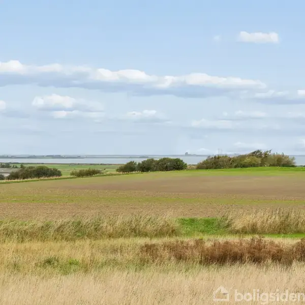 En vidstrakt mark ligger ubevægelig under en blå himmel med spredte skyer. Omgivet af lav vegetation og græsklædte områder, ses en fjern horisont mod havet. Inget tekst er synlig.