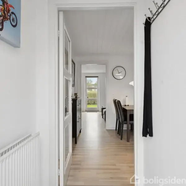 Et lyst indgangsparti fører ind til en stue med spisebord og stole. En vægdekoration med motorcykelmotiv hænger til venstre over en radiator. En vægur er synligt på væggen.