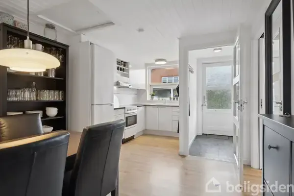 Et køkken med hvidt interiør, herunder skabe og et komfur. Et skab med glas og skåle står ved siden af et spisebord med stole. Udsigt mod en dør til baghaven.