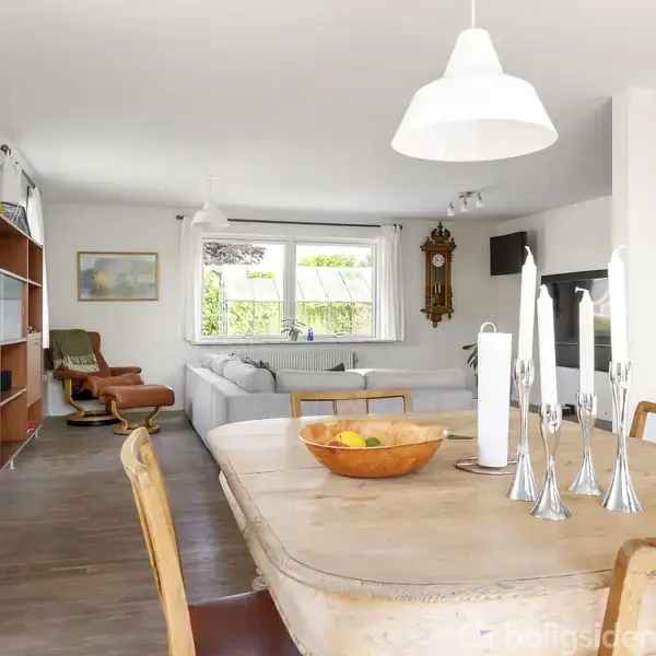 Et træbord er dækket med lysestager og en frugtskål, placeret i en lys stue med træmøbler, sofa og vindue, der giver udsigt til en grøn have.