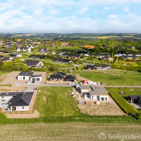 Boligområder beliggende midt i et frodigt, bakket landskab. Flere moderne huse og byggegrunde ses, omgivet af grønne marker og spredte træer under en blå himmel med skyer.