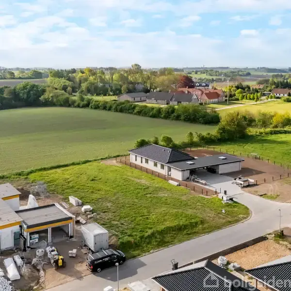 Et parcelhus ligger fredeligt øverst på en grusvej omgivet af marker og træer i et landligt område. Andre bygninger ses under opførelse i nærheden.