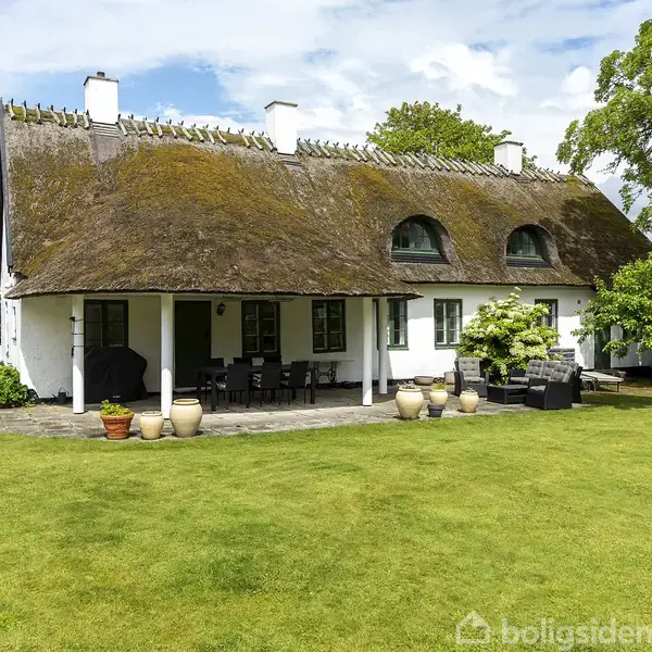 Stråtækt hus med hvid facade, placeret i en frodig have med grøn græsplæne. Huset har en udendørs terrasse med potteplanter og havemøbler under et overhæng.