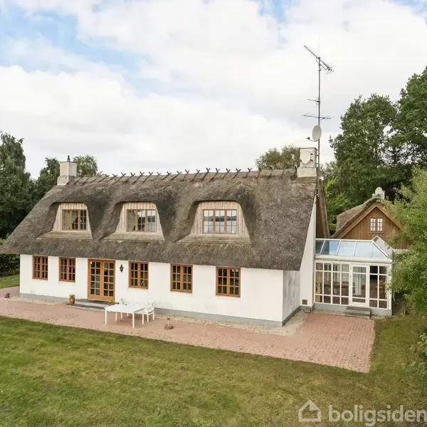 Stråtækt hus med hvide vægge og brunrammede vinduer, står på en flisebelagt terrasse. Omkringliggende græsplæne og skovklædt baggrund, med en tilbygget udestue på højre side.