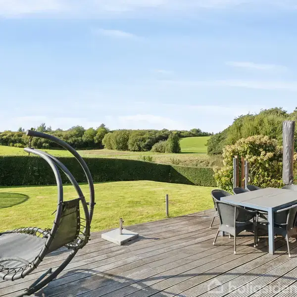 En hængestol og et havebord med stole står på en træterrasse. Udsigten viser en velplejet have med klippet græs og en hæk, omgivet af frodige grønne marker og skov.