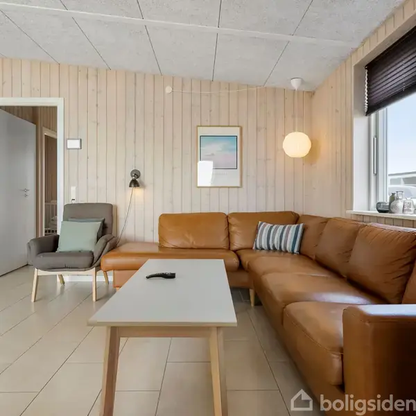 Brun lædersofa står i en lys stue med trægulv. Et lille bord foran sofaen bærer en fjernbetjening. Vindue med sort rullegardin og grøn plante i karmen giver udsigt udenfor.