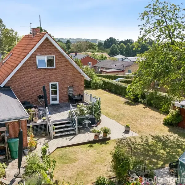 Et rødt murstenshus med sadeltag står i en velplejet have. Huset har en udendørs terrasse med trapper. Omkring ligger græsplæner, en hæk, et drivhus, og flere træer.