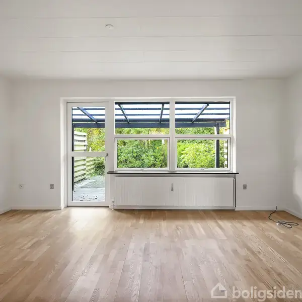 Et tomt rum med et trægulv fører til en terrasse gennem en glasdør. Store vinduer giver udsyn til grønne planter udenfor.