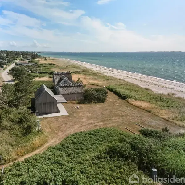 En række små træbygninger står langs en kystlinje ved havet, omgivet af grønne områder og en vej. Havet strækker sig til højre, med åben himmel i baggrunden.