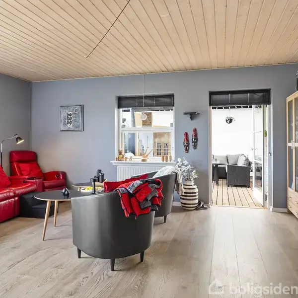 Stuen har røde lænestole, et gråt sofabord med dekorativ genstand, og en hvid trævæg. I baggrunden vises et vindue, dør til overdækket terrasse, lys trægulv og lofter.