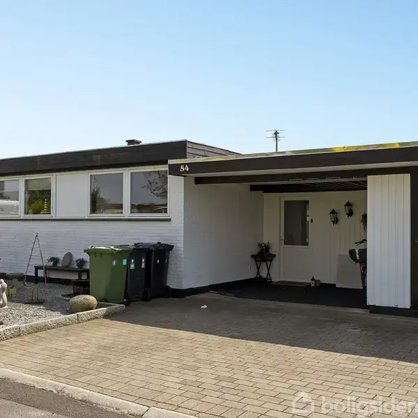En enkeltstående, hvidmurstenshus med to grønne affaldsbeholdere foran. Numret "84" vises ved siden af carporten. Omgivet af en flisebelagt indkørsel og haveudstyr på terrassen.