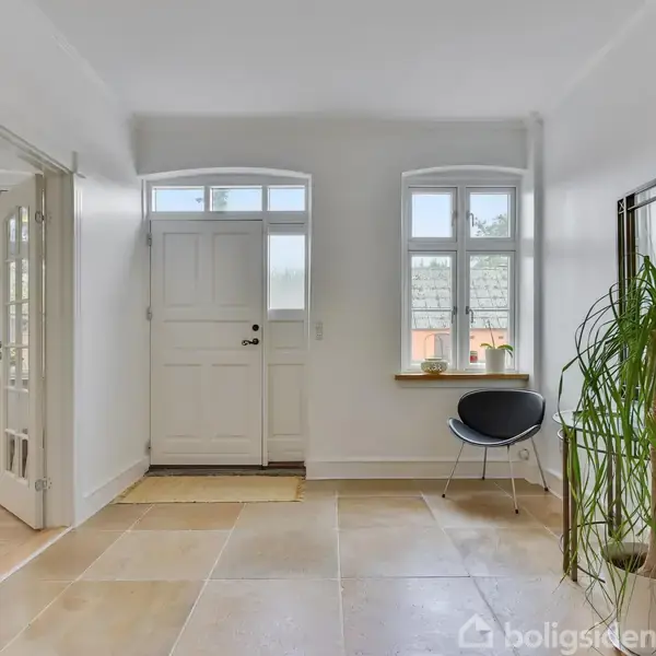 Entré med hvid dør og to vinduer, har store lyse fliser. Til højre er en stol og en plante, samt et spejl. Dør til venstre fører til andet rum.