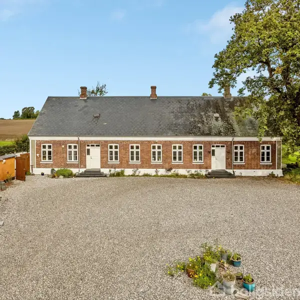 En lang murstensbygning med hvide vinduer og sort tag står stille på en grusbelagt gårdsplads. En gammel træbygning er til venstre, og et stort træ står til højre.
