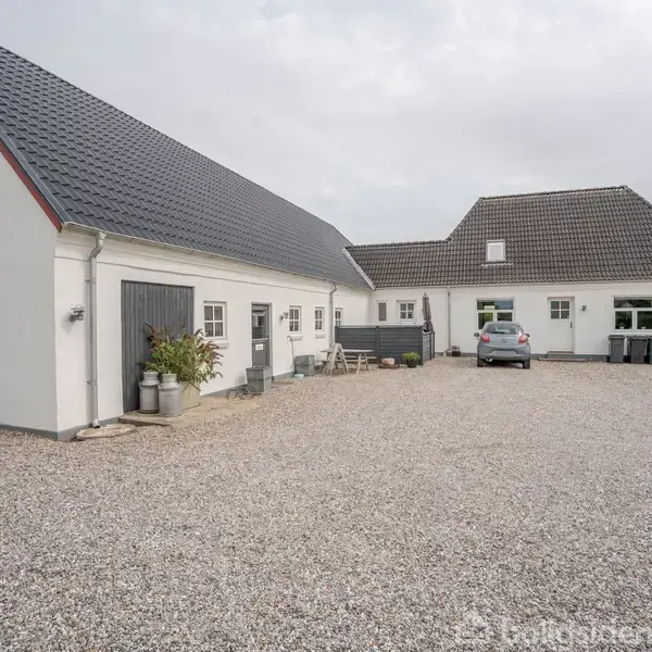 Et hvidt hus med gråt tag står stille på en grusbelagt gårdsplads. I baggrunden ses en parkeret bil og nogle grønne planter ved indgangen.