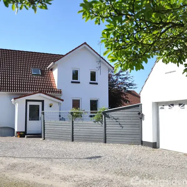 Et hvidt hus med rødt tegltag står ved siden af en garage; omgivet af en grusbelagt gårdsplads og træer i baggrunden.