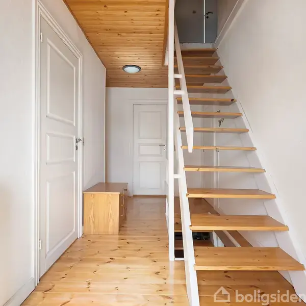 En trætrappe fører opad i en lys gang med trægulve og hvidmalede vægge. Til venstre er der en radiator og to hvide døre. En lille bænk står for enden.