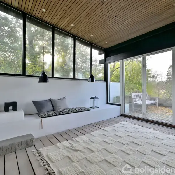 Skibenk med puder og lampe står i et skarpt, lyst rum med store vinduer og udsigt til en terrasse og natur. Tæppe på gulvet, "bo" dekorativt på hylden.