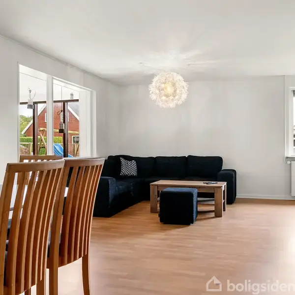 Svart sofa står i hjørnet af en stue med trægulv og et lille sofabord foran. En spisebord med træstole er til venstre, mens en rund lampe hænger fra loftet.