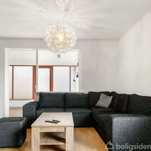 En mørkegrå sofa står i en stue med lyse trægulve, foran et rektangulært sofabord med en fjernbetjening. En moderne lampe hænger over bordet.