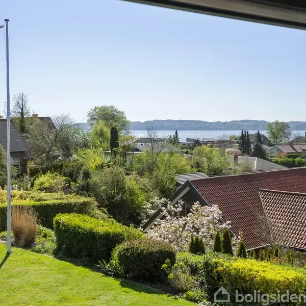 Flagstang med dannebrogsflag vajer i let vind. Omkringliggende haver med buske og træer, huse med røde tage ses. I baggrunden ses en blå fjord mod en klar himmel.
