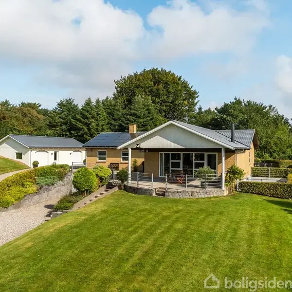 Hus med veranda står i solrig have med en velplejet græsplæne og omkranset af træer. En grusvej fører op til en garagebygning til venstre.