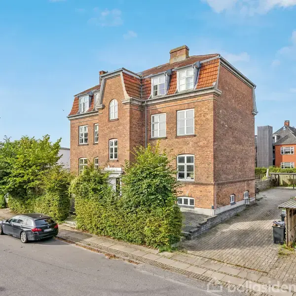 En stor, rød murstensbygning med tre etager og skrå tag. Bygningen har flere vinduer og omgives af buske og en parkeret bil. Beliggende på en stille villavej.