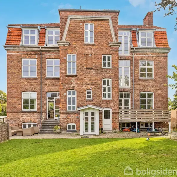 En stor, murstensbygning står med rødt tegltag, omgivet af en velholdt græsplæne. Der er fodboldmål og et skur i haven. Flot, blå himmel i baggrunden.