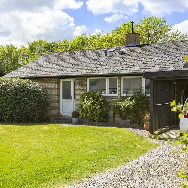 Et lille, enkelt hus med fladt tag og gule mursten står stille omgivet af en velplejet have med græs og buske. En bil holder delvist i en carport ved siden af huset.