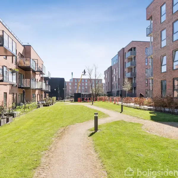 En stue med en grå sofa står langs væggen. Et rundt træbord er foran sofaen. Bagved er der store vinduer, der giver udsigt til en altan og en murstensbygning.