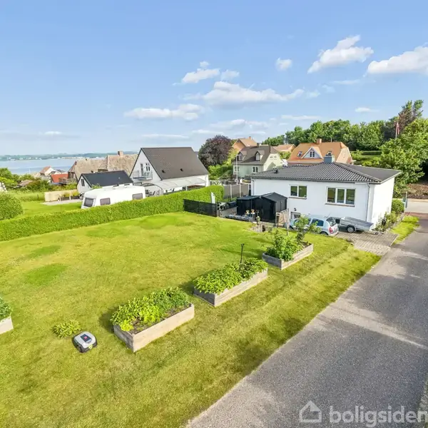 Hus med hvid facade omgivet af græsplæner og blomsterbede i en rolig beboelseskvarter. Vejen løber langs højre side, og flere huse ses i baggrunden under en klar blå himmel.