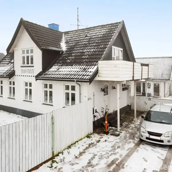 Et hvidt parcelhus med mørkt tag står på en snedækket grund. En bil er parkeret i indkørslen foran huset. Omkringliggende hegn omkranser ejendommen.