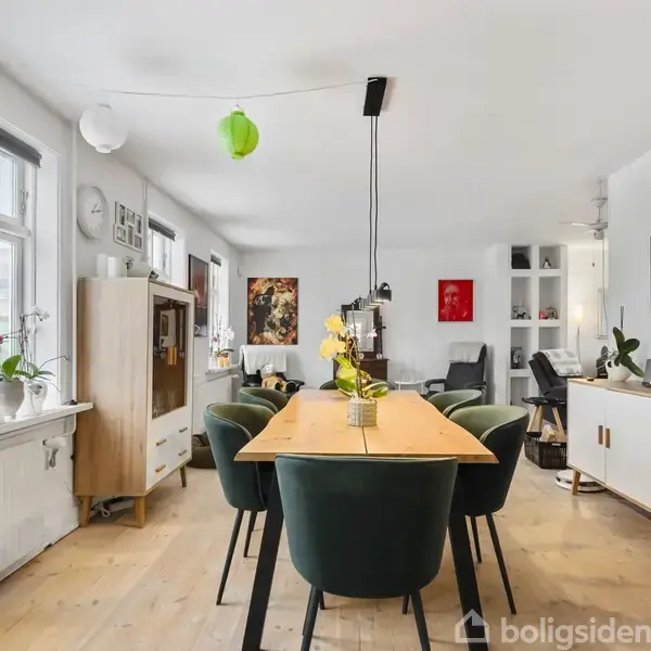 En lys spisestue med et træbord omgivet af grønne stole. En vase med blomster står på bordet. Rummet indeholder også skænke, planter og kunstværker på væggene.