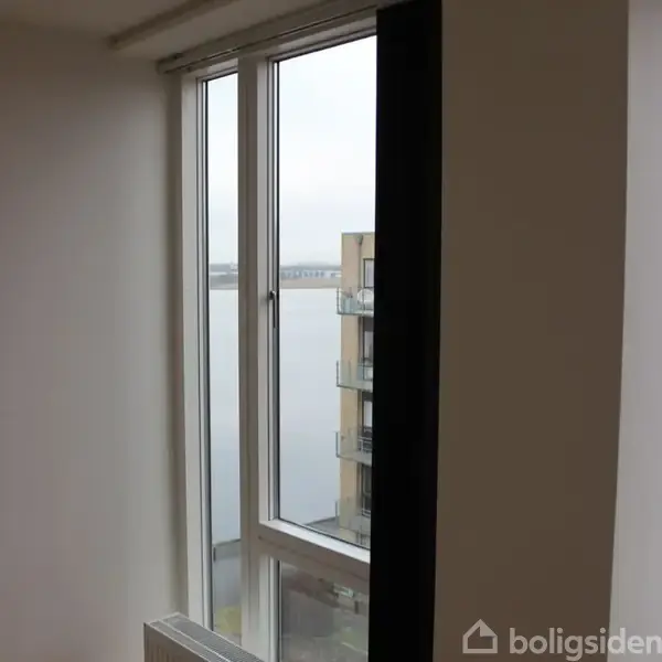 Et vindue åbner ud mod en udsigt over en del af en bygning med altaner og en stor vandflade i baggrunden under en overskyet himmel.