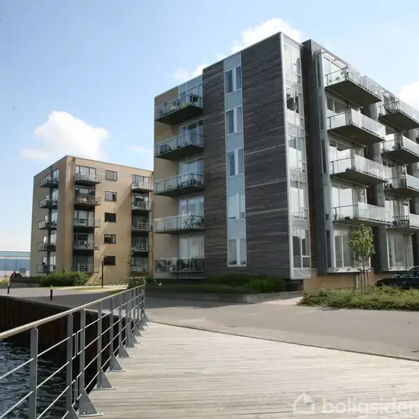 Et moderne lejlighedskompleks med flere etager står langs en havnefront. En flisebelagt gangsti og parkering løber ved siden af bygningerne. Himlen er delvist overskyet.