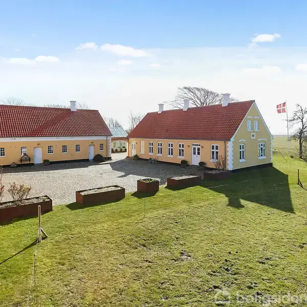To gule bygninger med røde tage står på en grusplads omgivet af en grøn plæne. Dannebrog vajer på en flagstang. Skoven ses i baggrunden.