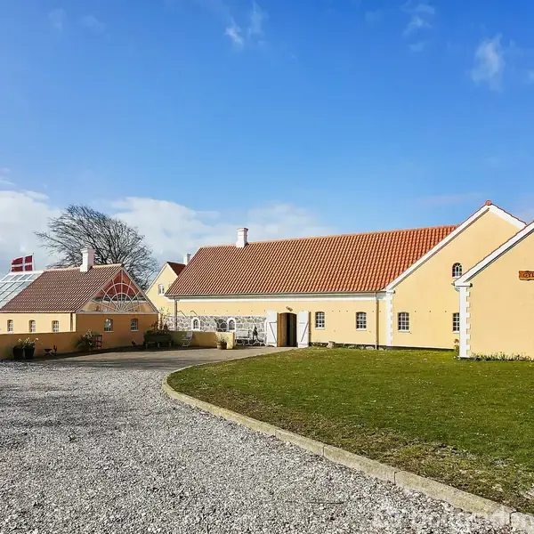 Et gult bondehus med rødt tegltag står på en grusvej omgivet af en grøn græsplæne. Et drivhus med Dannebrog er til venstre.