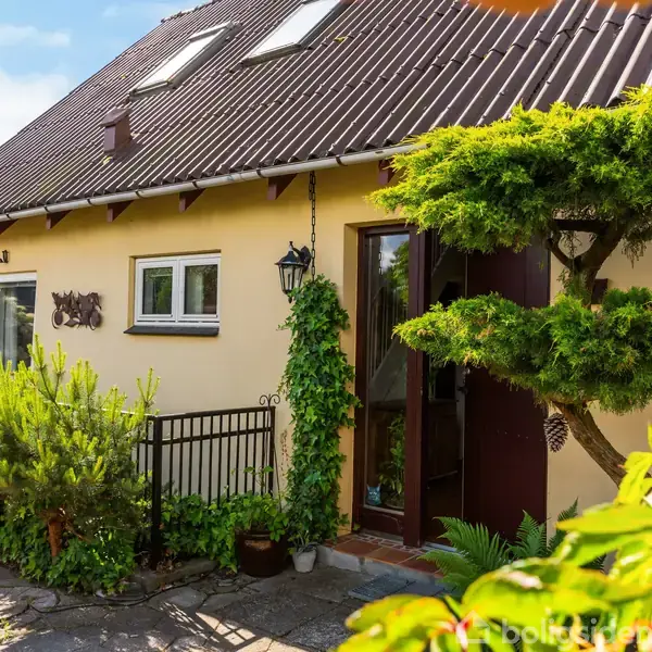 Et gulmalet hus med skråt tag ses, omgivet af grønne planter og buske. En lanternestylte hænger ved siden af hoveddøren, og terrassen er omgivet af frodig vegetation.