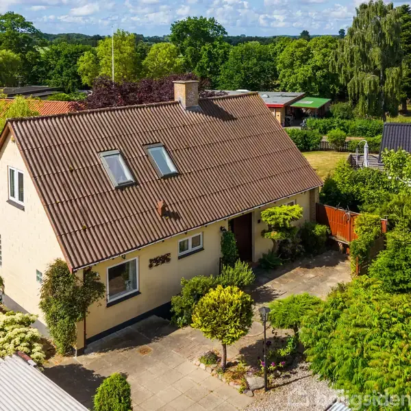 Et gult murstenshus med brunt tegltag står omgivet af en frodig have. Huset har tagvinduer og en indgang beliggende mellem grønne planter. Baggrunden består af tæt beplantning og flere huse.