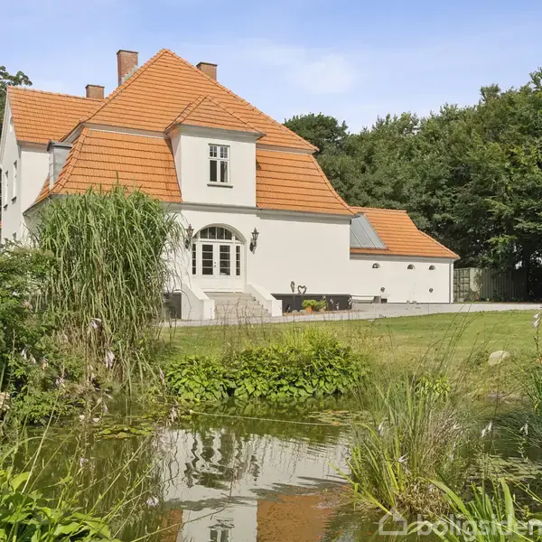 En stor hvid villa med orange tegltag står omgivet af en grøn have med en lille dam i forgrunden, omgivet af buske og blomster, træer i baggrunden.