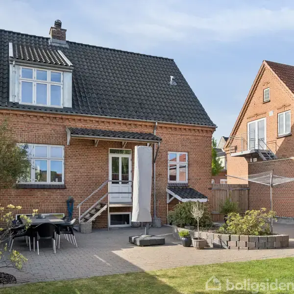 Et rødt murstenshus med sort tegltag i en baghave. En terrasse med havemøbler og planter omringet af en træhegn, under en blå himmel.
