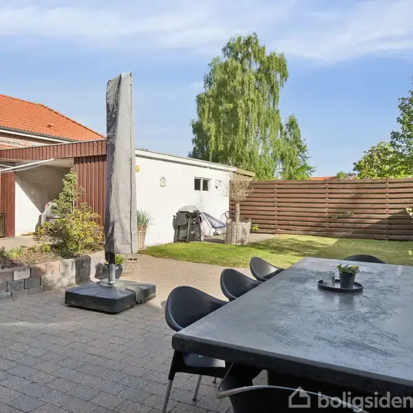 Terrasse med stort spisebord og stole i en flisebelagt gårdhave. I baggrunden ses en lille græsplæne, et hegn, og en hvid bygning i et solrigt miljø.