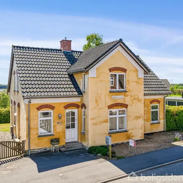 Gult hus med sort tegltag står stille ved vejen, omgivet af grønne buske og himmel i baggrunden. Murede detaljer omkring vinduer. Formiddagssol belyser facaden.